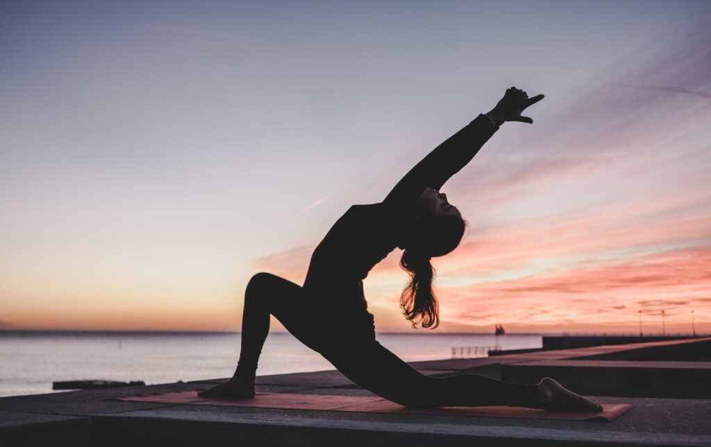 Women practicing yoga on a mat outdoors near water at sunset with colorful sky.