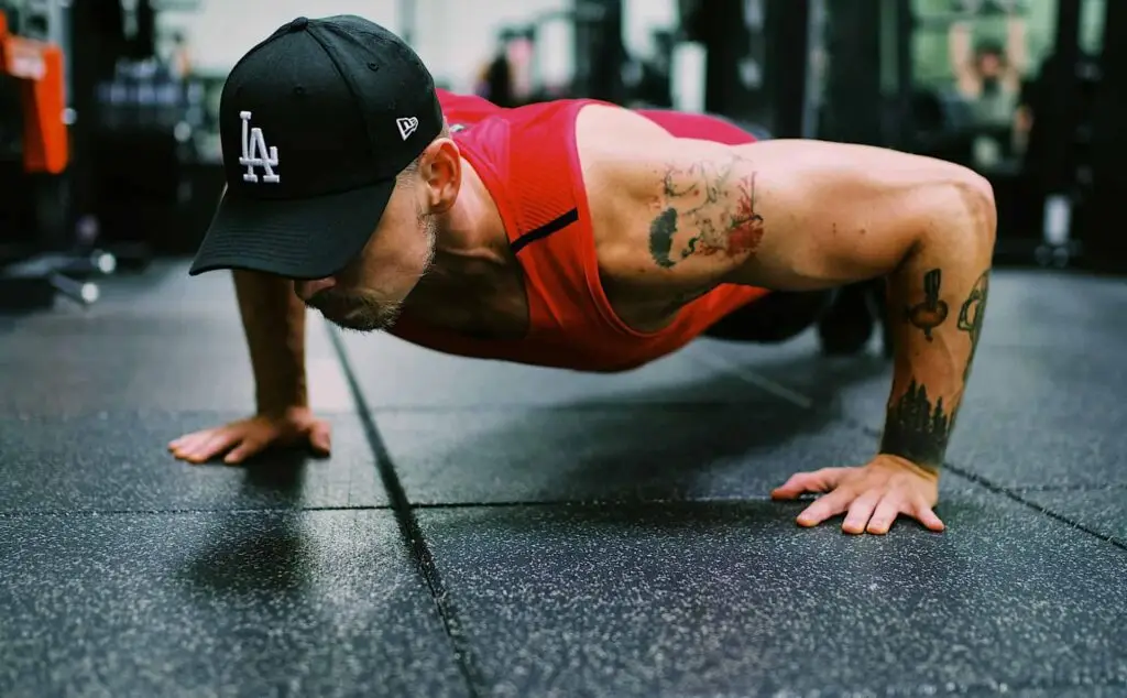 Man in black cap and red sleeveless shirt doing a push-up on gym floor with arm tattoos.
